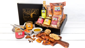 A black gift box with gold embossed tree with a frog alongside brightly colored jars of fruit conserves and dried fruit, and handmade cookies 