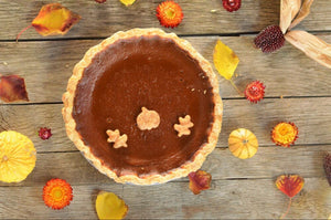 A flatlay image of a golden brown pumpkin pie, sitting in a buttery crust, surrounded by fall leaves and flowers for decoration. 