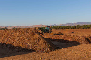 A wide image of 2 large piles of brown compost in the foreground, with a tractor in the background, against a blue sky.