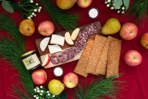A flat lay with a deep red table cloth spread with green pears, pink apples, slices of lemon salami, and triple cream brie cheese with holiday greenery. 