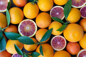 A close up image of bright and whole blood oranges, with vibrant green leaves and halves of juicy red blood oranges intermixed.