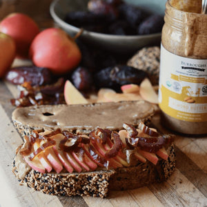 An open faced toast with almond butter, apple slices, and dates, sitting atop a wooden counter with the ingredients in the background.