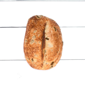 A close up flatlay of a rustic, golden loaf of bread with bits of dark olives in it, set against a white wooden background.