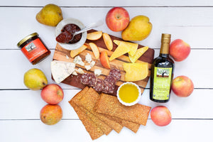 A flat lay featuring a variety of cheese, crackers, olive oil, salami, jelly, and delicious apples and pears, artfully arranged on a white table.