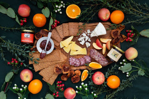 A flat lay featuring a variety of cheese, crackers, salami, jelly and marmalade, and delicious organic fruit, artfully arranged on a cloth with green leaves surrounding it.