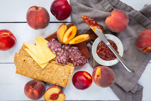 A flat lay featuring a variety of cheese, crackers, olive oil, salami, jelly, and juicy peaches and nectarines, artfully arranged on a white table and wooden serving board.