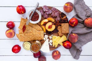A flat lay featuring a variety of cheese, crackers, salami, jelly and marmalade, and juicy peaches and nectarines, artfully arranged on a wooden serving board.