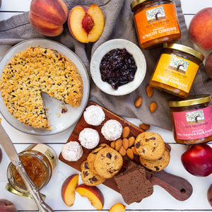 An artful spread of an assortment of handmade jams, baked goods, nuts, surrounded by juicy peaches and nectarines.