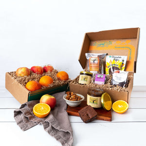 Two rustic craft gift baskets featuring vibrant oranges and crisp apples, and artfully packaged granola, marmalade, brownie, tea, and chocolate covered pistachios.