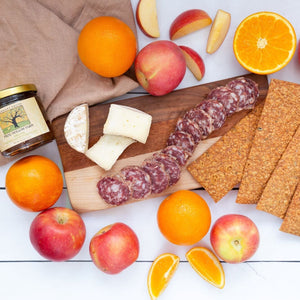 A flat lay of sliced salumi, creamy brie style cheese, a jar of handmade marmalade and artisan crackers displayed on a wooden cutting board against  white wooden background. 