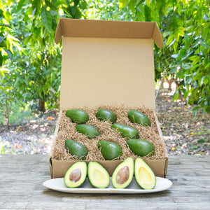 An image of a rustic, craft gift basket with light green, buttery avocados on a wooden table, set against a background of greenery.