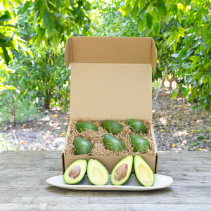 An image of a rustic, craft gift basket with light green, buttery avocados on a wooden table, set against a background of greenery.