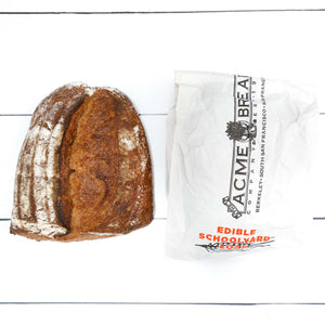 A closeup of a loaf of rustic, artisan bread with a crunchy golden crust, next to a white bag that says "Edible Schoolyard Loaf" on it.