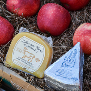A close up image of fresh, crisp apples next to a packaged Bellwether Farms Carmody Cheese and a Point Reyes Original Blue Cheese, in a rustic craft gift basket.