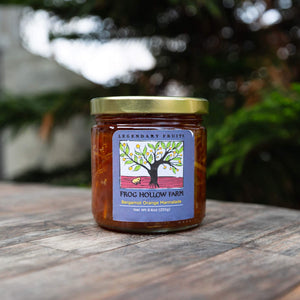 A jar of bergamot orange marmalade with a colorful Frog Hollow label, sitting on a rustic wooden table with trees in the background.
