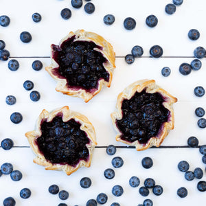 A flat lay featuring three golden pastry tartlets, filled with delicious juicy blueberries, surrounded by fresh blueberries.