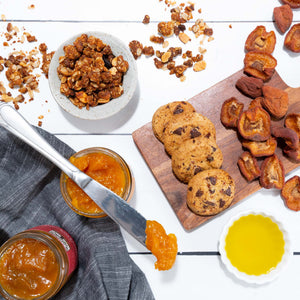 A flat lay image featuring handmade cookies, dried fruit, granola, olive oil, and fruit conserves artfully arranged in dishes. 