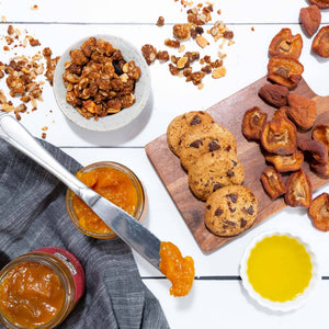 A flat lay image featuring handmade cookies, dried fruit, granola, olive oil, and fruit conserves artfully arranged in dishes. 