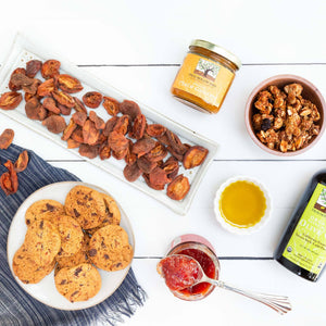 A flat lay image featuring handmade cookies, dried fruit, granola, olive oil, and fruit conserves artfully arranged in separate dishes.