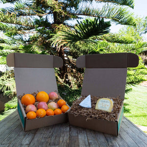 Two rustic craft gift baskets sitting atop a wooden table, one filled with a variety of colorful, fresh fruit and the other filled with packaged Bellwether Farms Carmody Cheese and Point Reyes Original Blue Cheese.