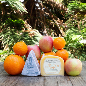 An image of Bellwether Farms Carmody Cheese and Point Reyes Blue Cheese, surrounded by a variety of vibrant, fresh fruit, sitting atop a rustic wooden table against a background of sunlit greenery.