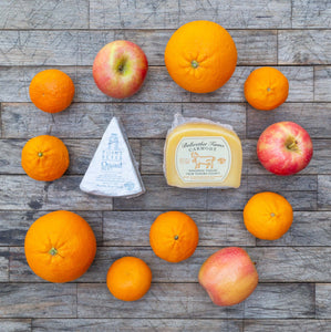 A flatlay image of Bellwether Farms Carmody Cheese and Point Reyes Blue Cheese, surrounded by a variety of vibrant, fresh fruit, sitting atop a rustic wooden table.