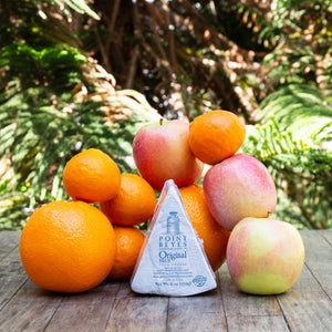 An image of Point Reyes Original Blue Cheese, surrounded by a variety of vibrant, fresh fruit, sitting atop a rustic wooden table against a background of sunlit greenery.
