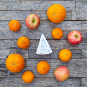 A flatlay image of a block of packaged Point Reyes Original Blue Cheese, surrounded by a variety of vibrant, fresh fruit, sitting atop a rustic wooden table.
