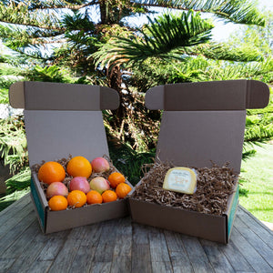Two rustic craft gift baskets sitting atop a wooden table, one filled with a variety of colorful, fresh fruit and the other filled with packaged Bellwether Farms Carmody Cheese.