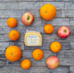 A flatlay image of a block of packaged Bellwether Farms Carmody Cheese, surrounded by a variety of vibrant, fresh fruit, sitting atop a rustic wooden table.