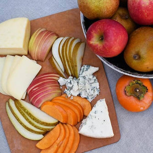 An artful spread of various fresh fruit slices, including apples, pears, and persimmons, next to blocks of fresh carmody and blue cheese. These ingredients are sitting atop a wooden serving board, next to a bowl of whole, fresh fruit.