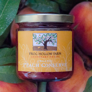 A flatlay of a jar full of Frog Hollow's organic peach conserve, on a background of fresh peaches and green leaves.