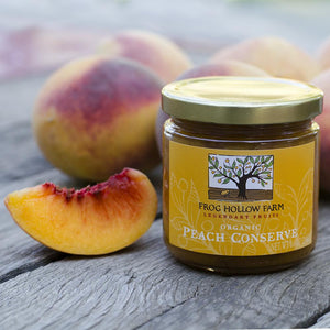A jar full of Frog Hollow's organic peach conserve, surrounded by fresh, juicy peaches on a rustic wooden table.