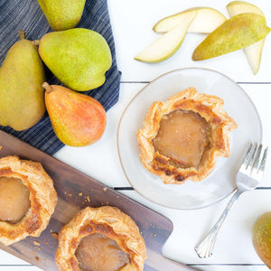 A flatlay of handmade pear frangipane tartlets, cradled in golden pastry, surrounded by fresh juicy pears on a white table.