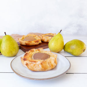 An image featuring three plated pear frangipane tartlets, cradled in golden pastry, surrounded by fresh juicy pears on a table.