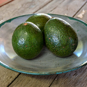 A close up image of three avocados with dark, bumpy skin, sitting in a bowl on a rustic wooden table.
